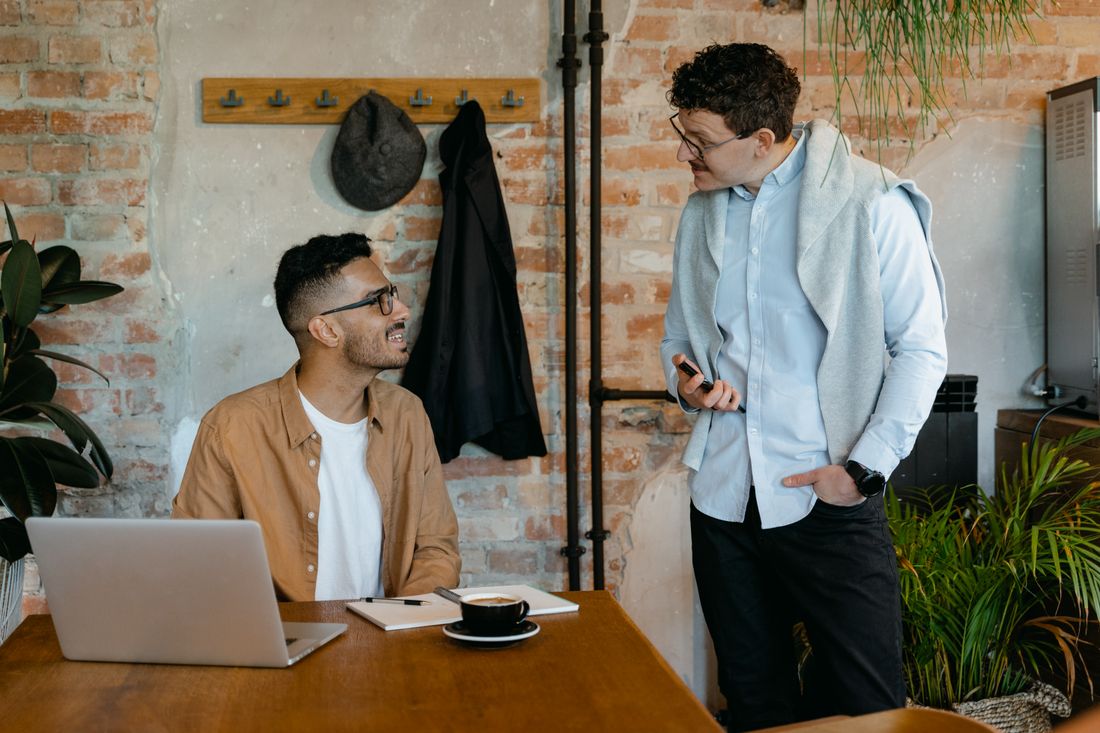 Two colleagues talking in a startup office with exposed brick walls, one in a casual layered jacket and one in a button-down shirt