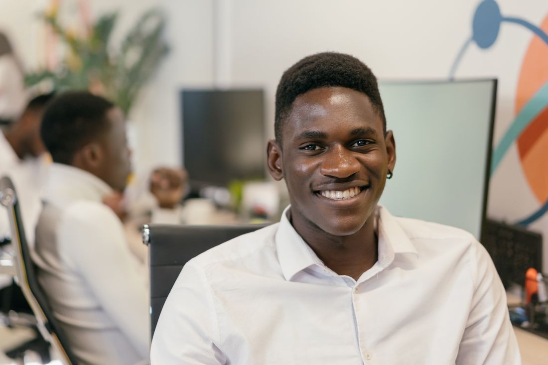 Man in a white button-down shirt smiling naturally at his desk in a modern office with a colleague visible in the background