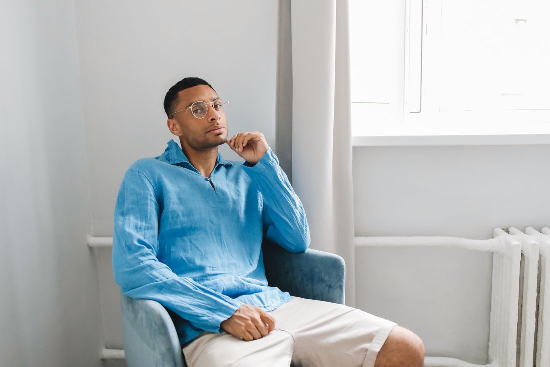 A man in a blue linen shirt and shorts sits in an armchair by a window, chin resting on his hand, looking off to the side.