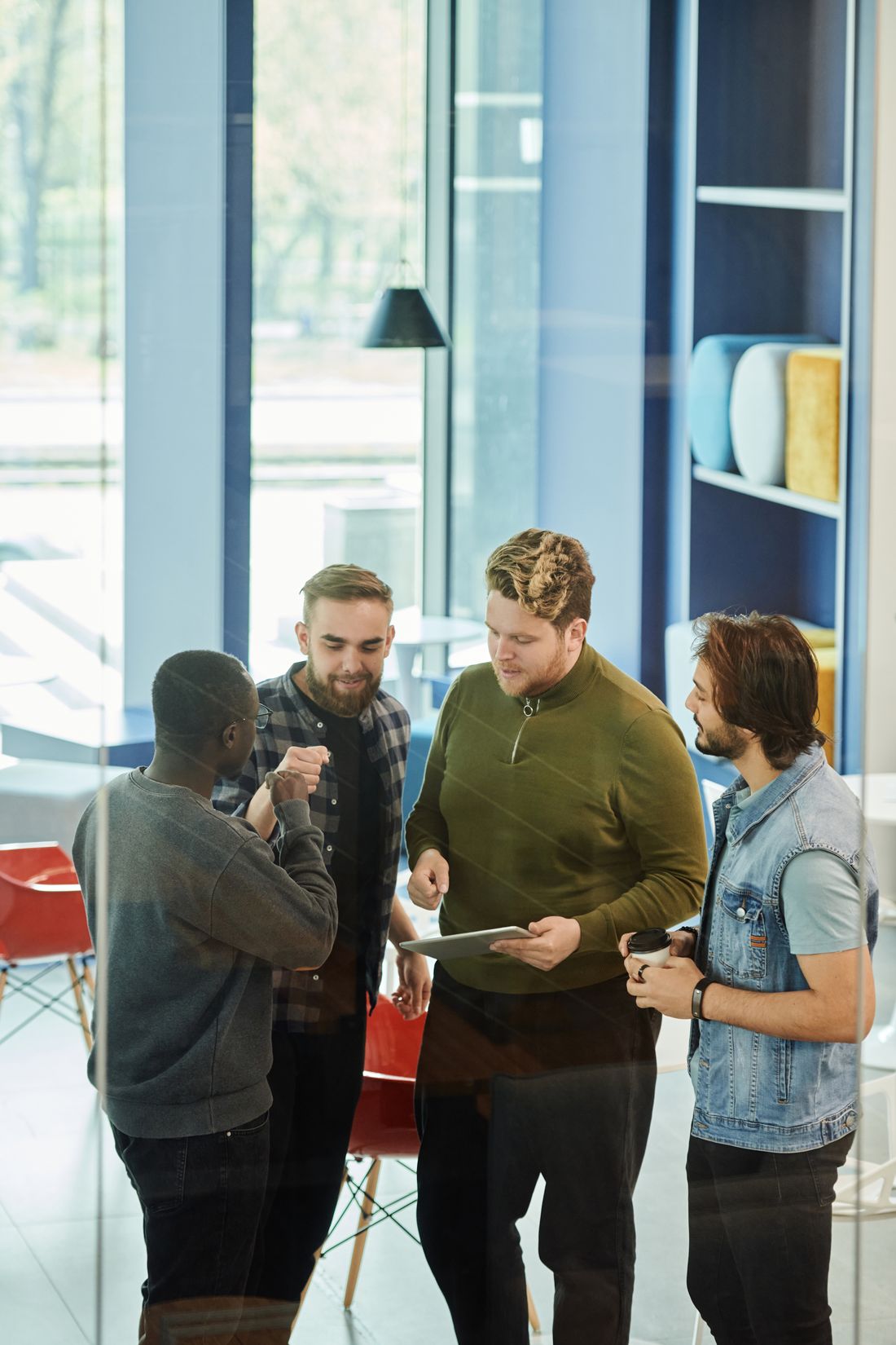 A small group of business-professional men standing together in a modern common area, looking at a tablet and talking.