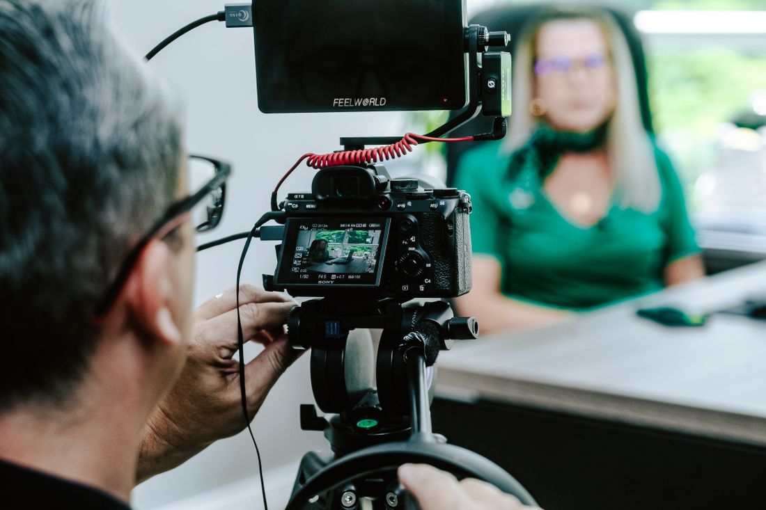 A camera operator monitors a subject on a small rig during a video interview in a naturally lit office setting.