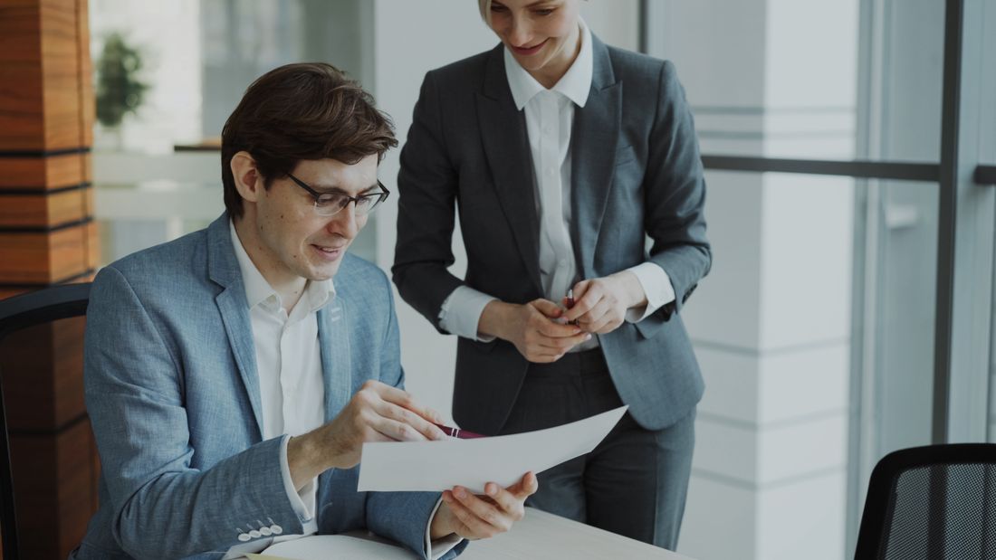 Two colleagues review a printed document together at a desk in a modern office.