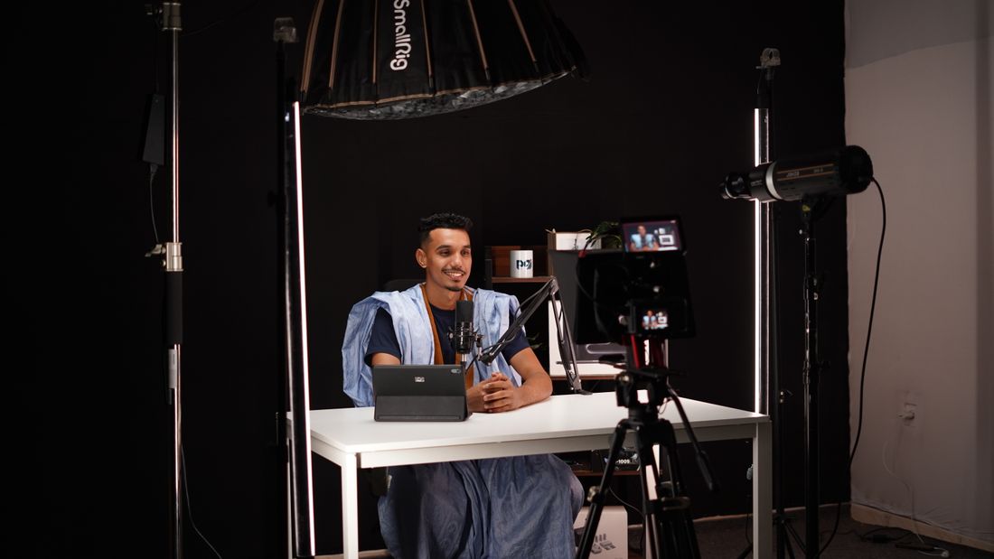 A man sitting at a small desk being filmed on camera under an overhead softbox light in a compact video studio setup