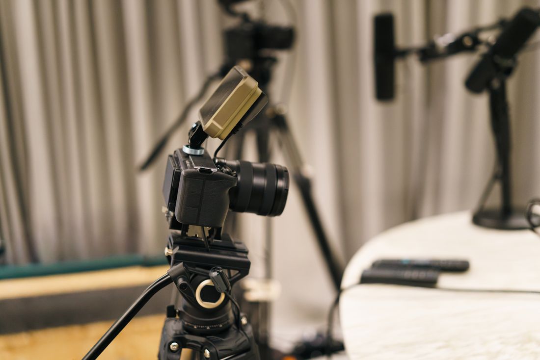 Close-up of a mirrorless camera mounted on a tripod with a small light attachment, framed against a curtain backdrop with a microphone in the background