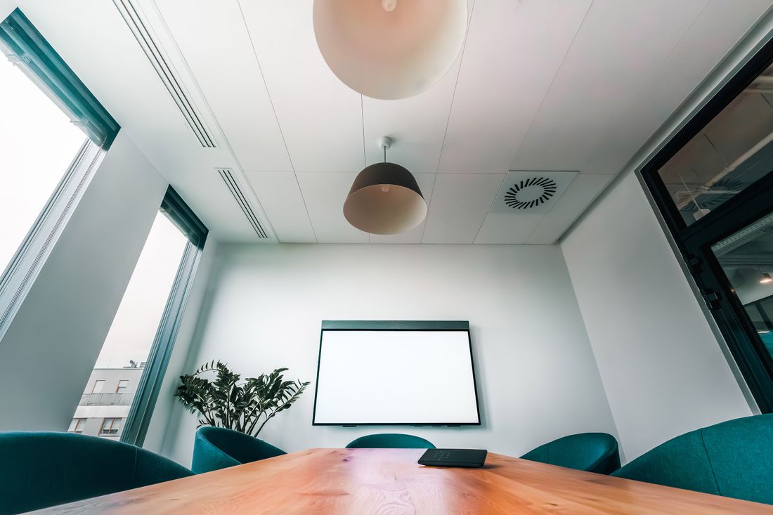 An empty, modern conference room with a blank wall-mounted screen, a wooden table, and a potted plant, set up ahead of a meeting or event.