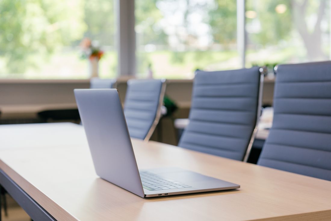 A laptop open on a wooden conference table in an empty room, with rows of chairs visible in the background.