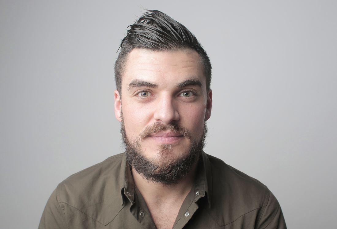 A bearded man in a plain olive shirt photographed head-and-shoulders against an even grey background.