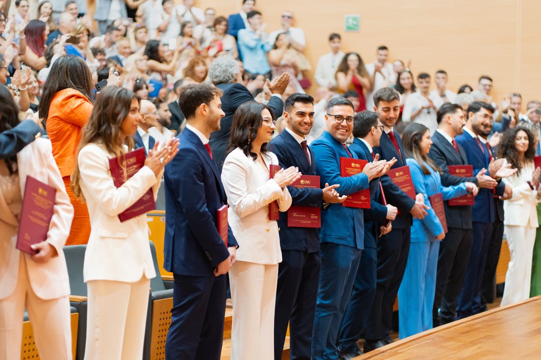 Graduates and guests applaud during a formal indoor university ceremony, rows of red diploma folders visible in the crowd