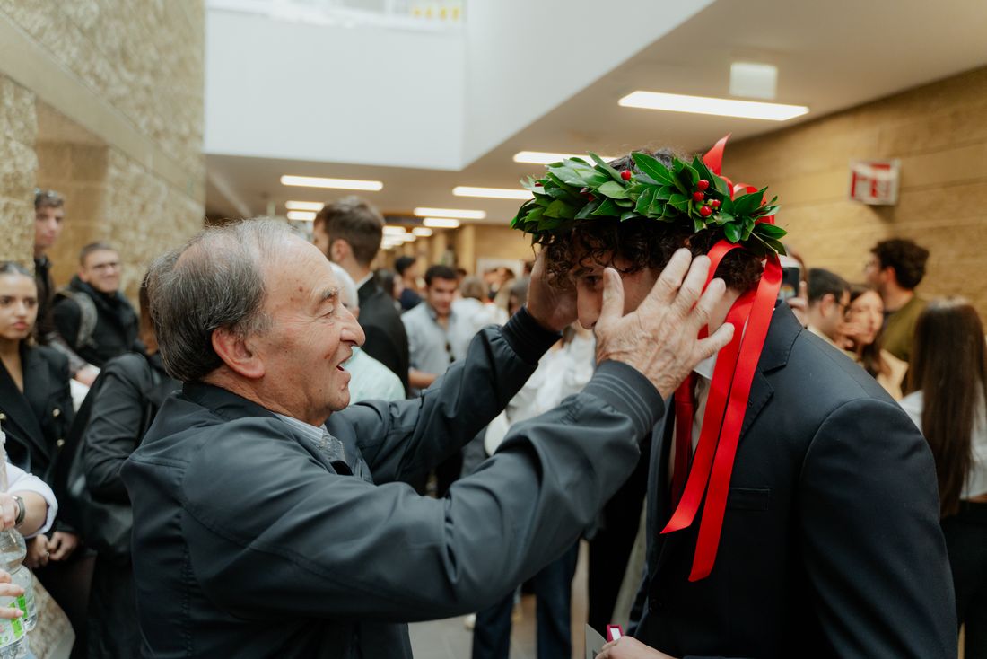 An older man places a laurel wreath on a younger graduate's head in a candid, unposed moment in a campus hallway