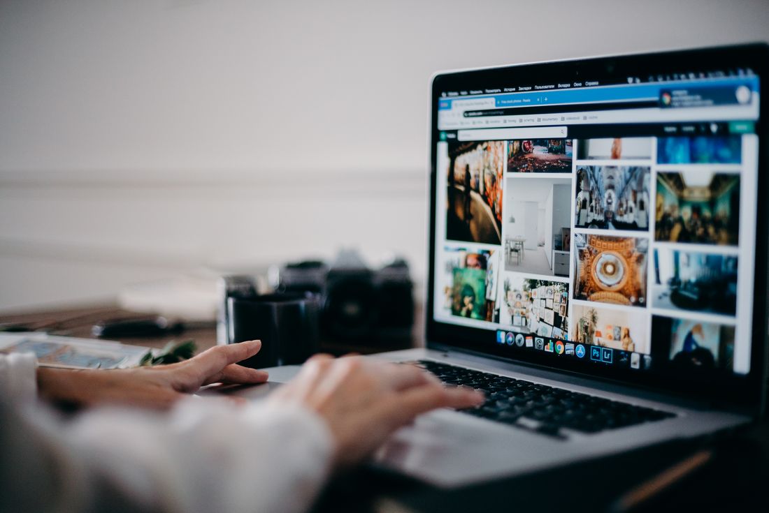 A person's hands typing on a laptop showing a grid of thumbnail photos on screen, with a camera resting nearby on the desk