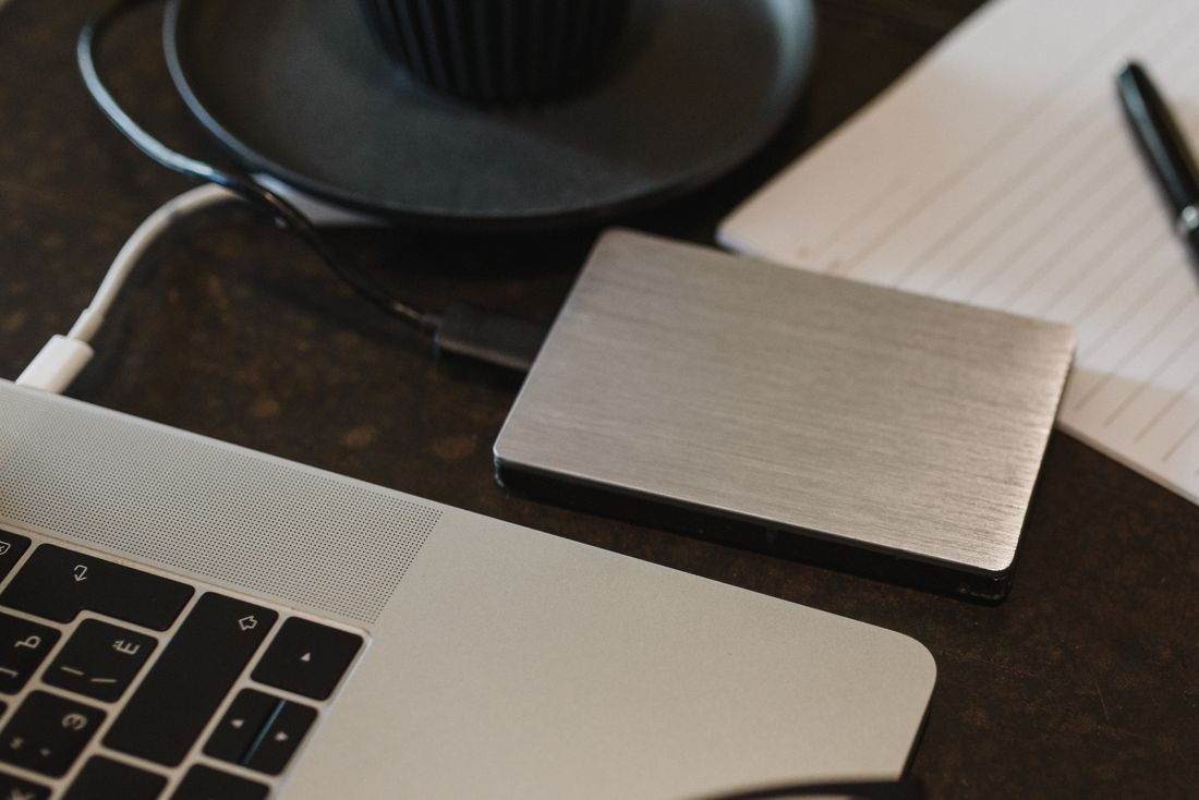 Portable external drive connected to a laptop beside a notepad and pen on a dark wood desk