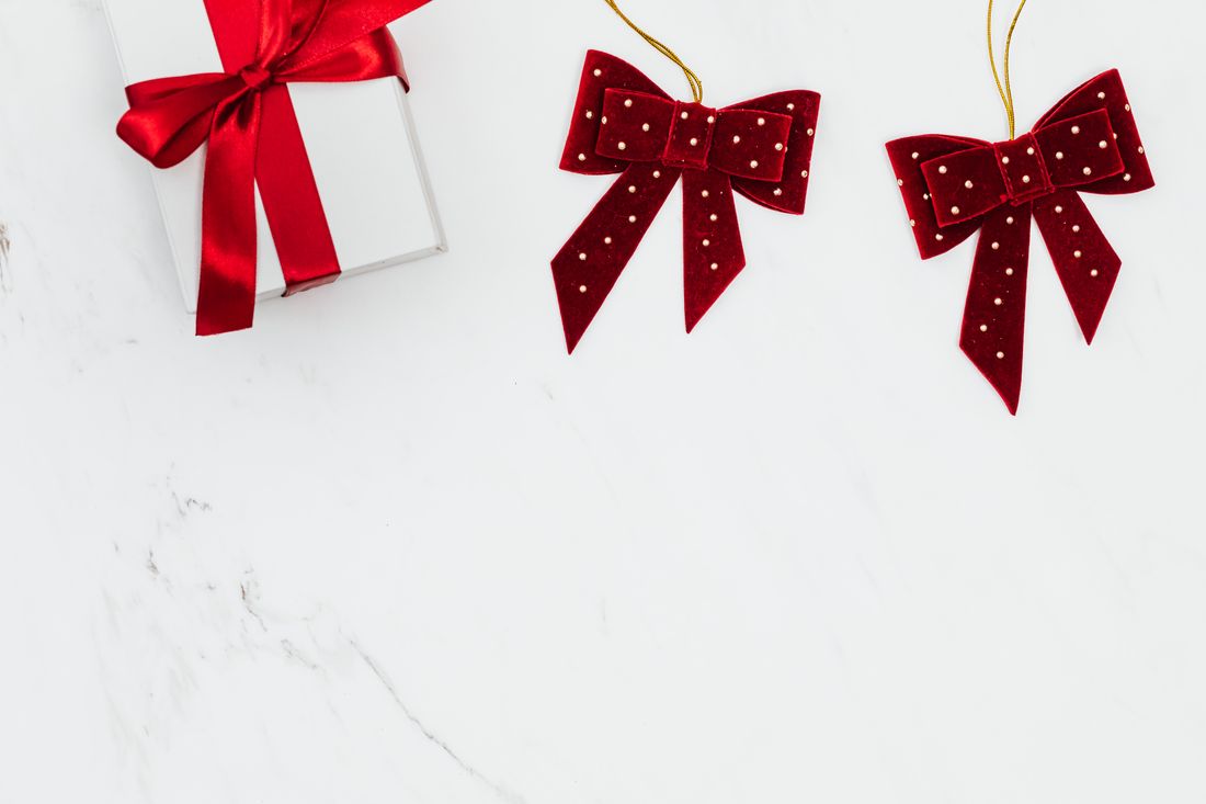 A red ribbon-tied gift box and two red velvet bows laid flat on a plain white marble surface.