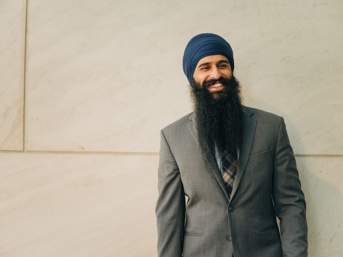 A smiling professional in a grey suit and patterned tie standing against a plain stone wall in daylight.