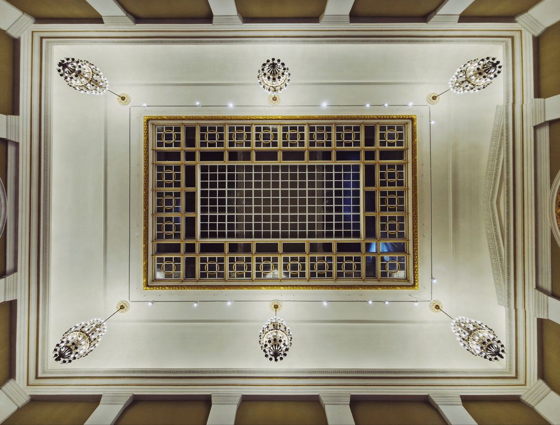 Ornate ceiling seen from below, with a gilded grid skylight panel and small chandeliers at each corner