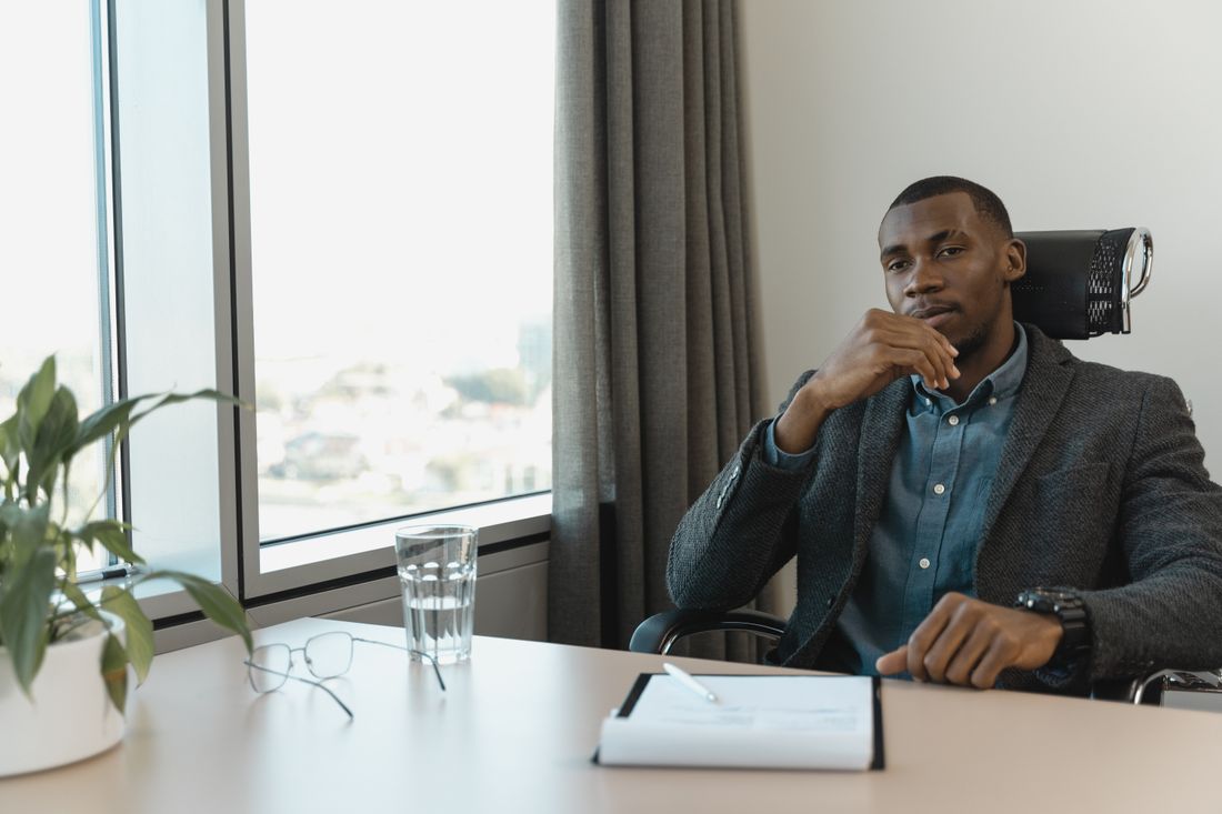 A businessman sits thoughtfully at a clean desk near a window with a city view, holding his chin, with a closed notebook, glasses, and a glass of water on the desk.