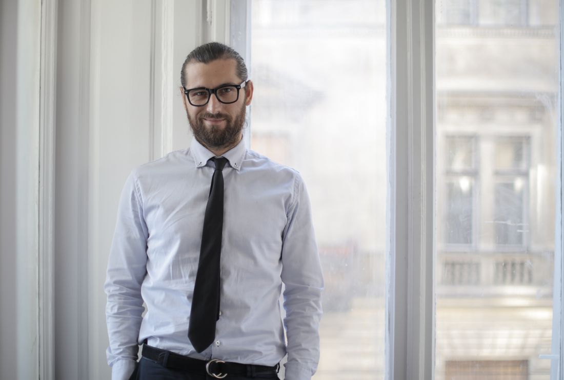 A professional in a light dress shirt and tie stands calmly near a bright window in a plain interior, looking off to the side.