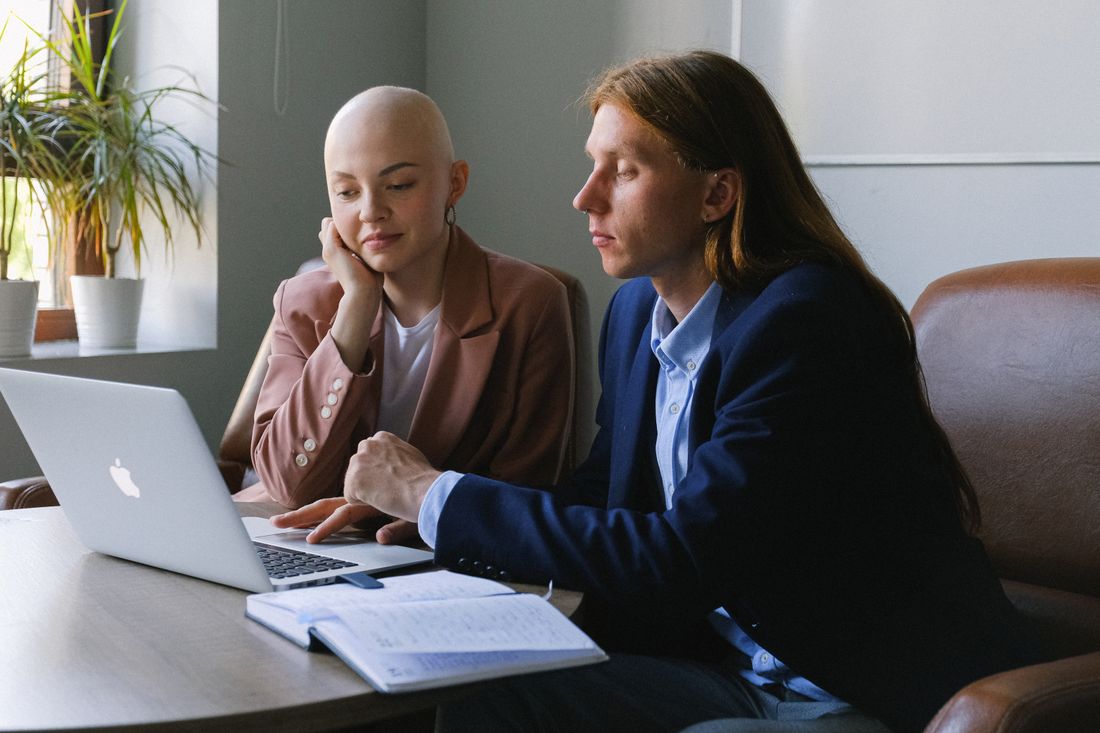 Two colleagues in business attire reviewing a laptop screen together at a table with a notebook open, in a bright office.