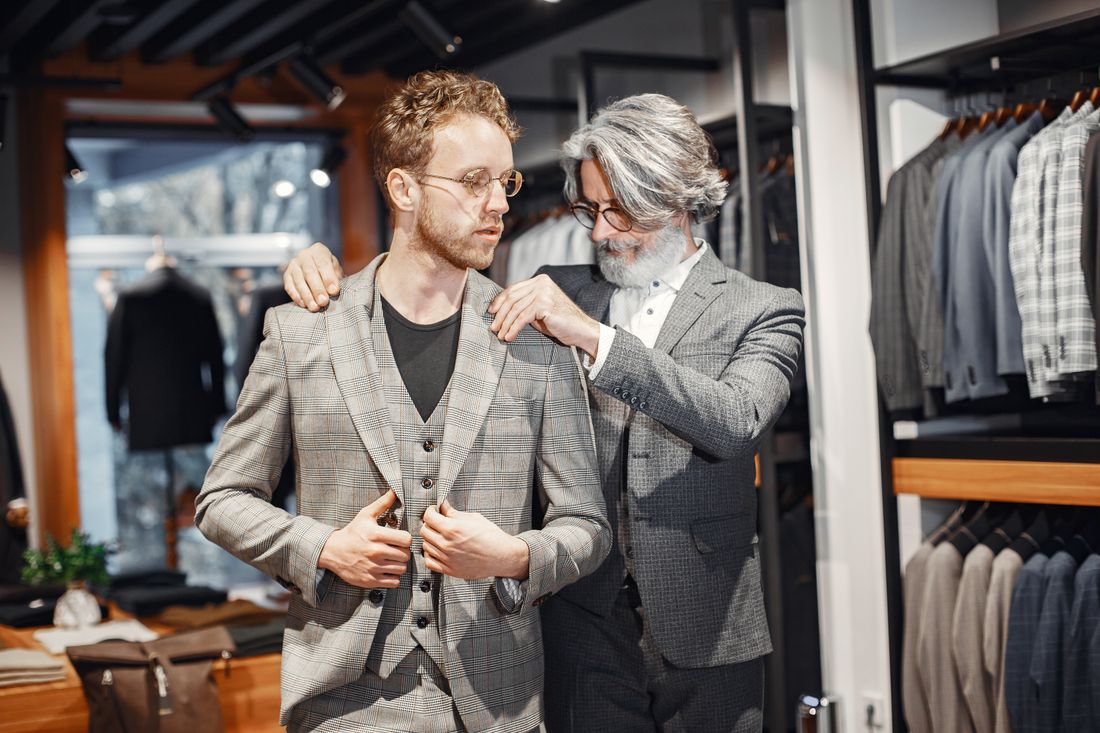 An older stylist adjusts the shoulders and collar of a plaid jacket on a younger man in a boutique clothing shop.