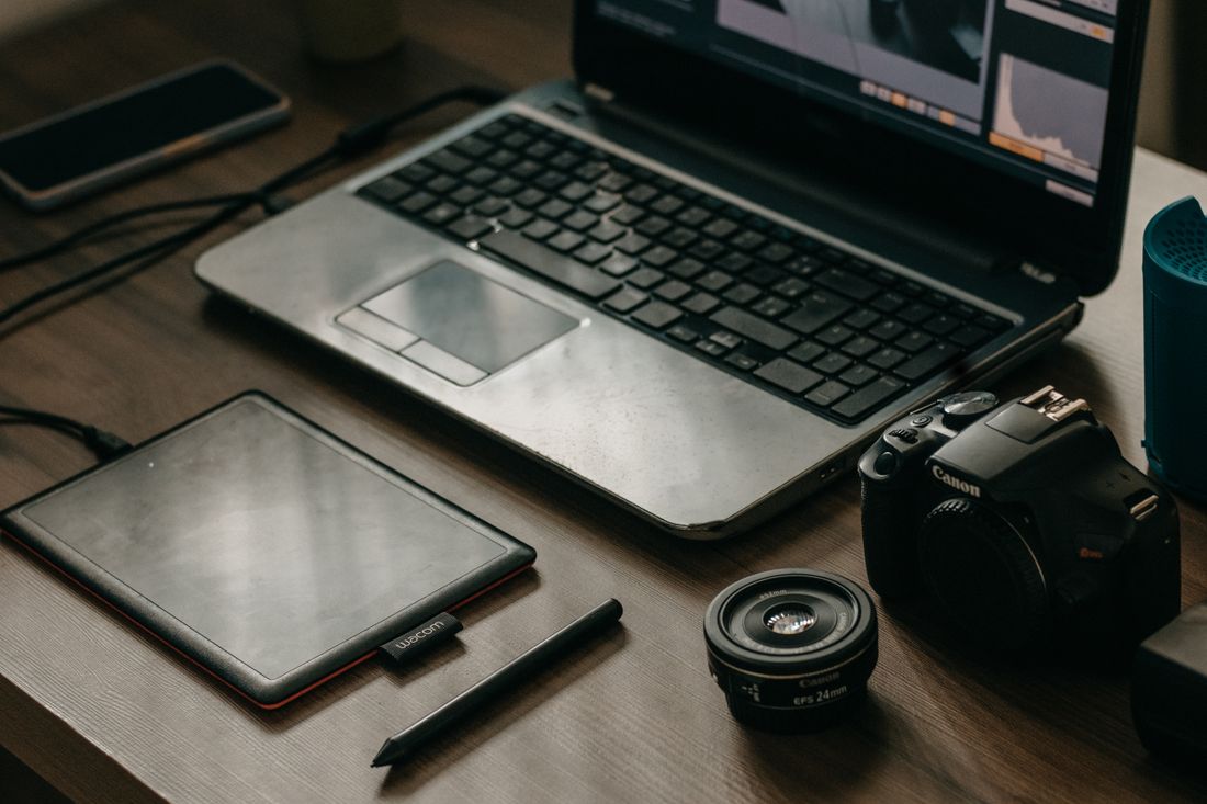 A laptop, camera body, lens, and graphic tablet arranged on a desk, representing the photo and video assets a website redesign has to produce.