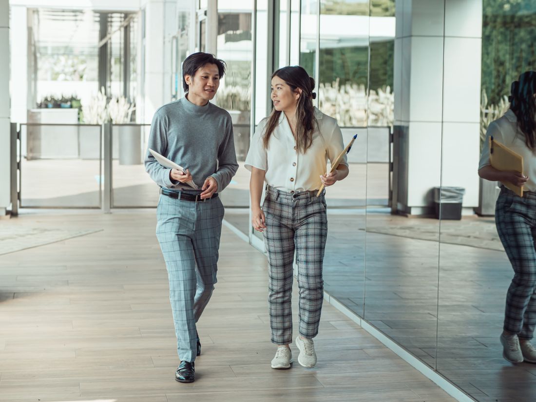 Two colleagues in conversation while walking through a bright office corridor, holding notebooks, mid-distance with faces not the focus.