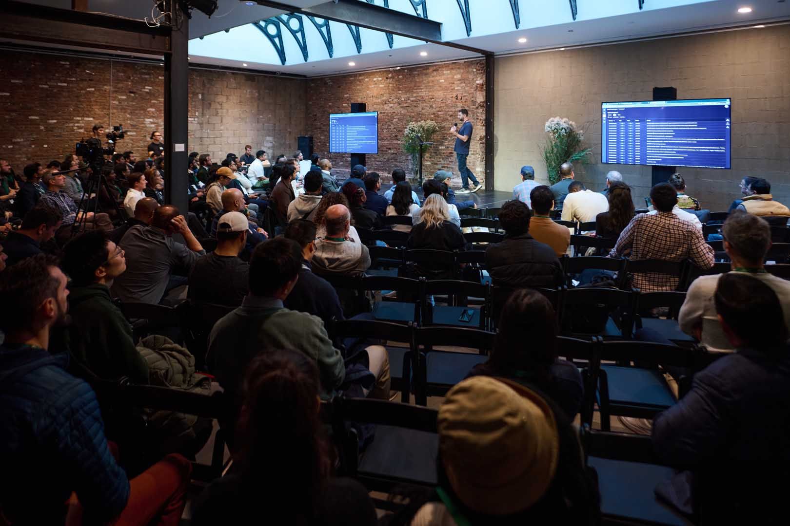 A seated audience in a brick-walled venue faces a speaker between two screens, with video cameras on tripods set up along the side of the room.