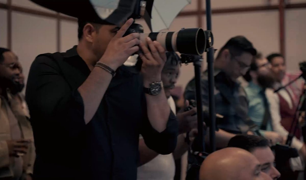 A photographer shoots with a long lens from the aisle of a seated event audience, a light stand beside him.