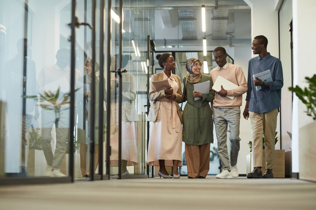 A group of colleagues in business attire walk and talk together through a modern glass-walled office hallway.
