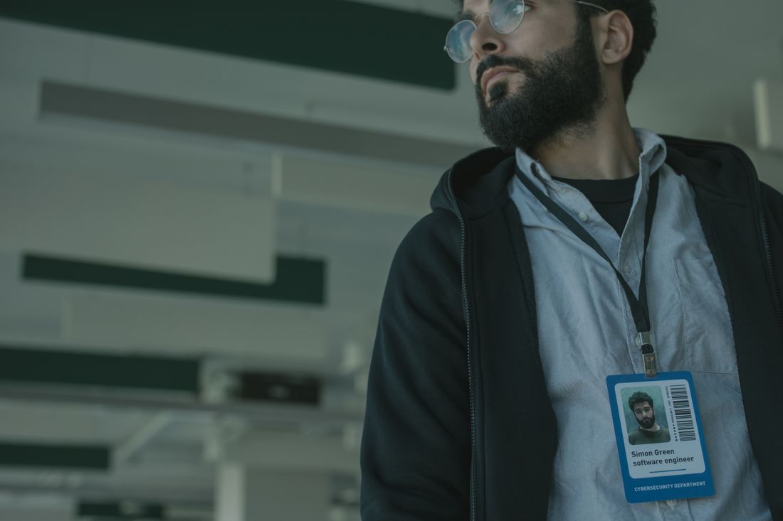 A man wears glasses and an ID badge with a printed photo on a lanyard in a modern office.