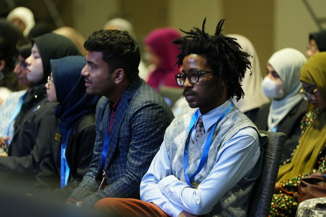 A diverse audience of people in business attire seated and facing forward, attentively watching at an indoor conference.
