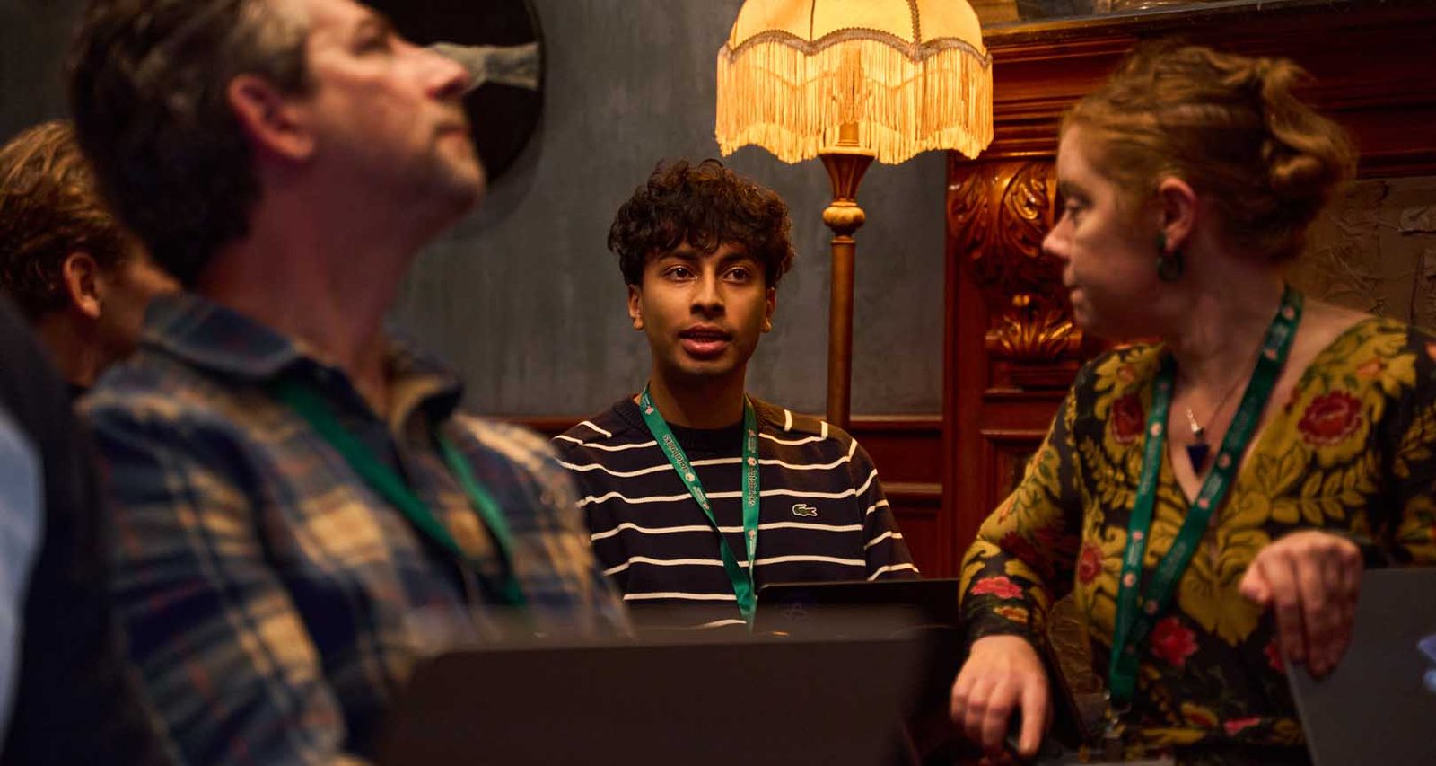 Three conference attendees talking under a warm table lamp at a Manhattan venue, event badges visible, shot candidly in low light