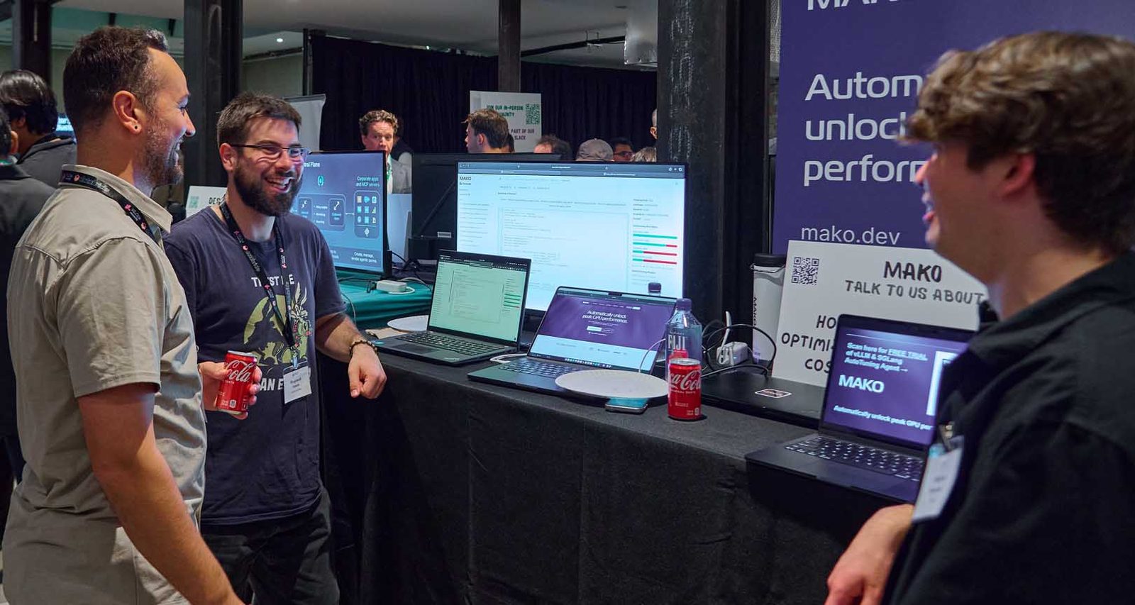 Three attendees talking at a sponsor booth at an NYC tech conference, with a branded backdrop, laptops, and a scan-here QR sign behind them