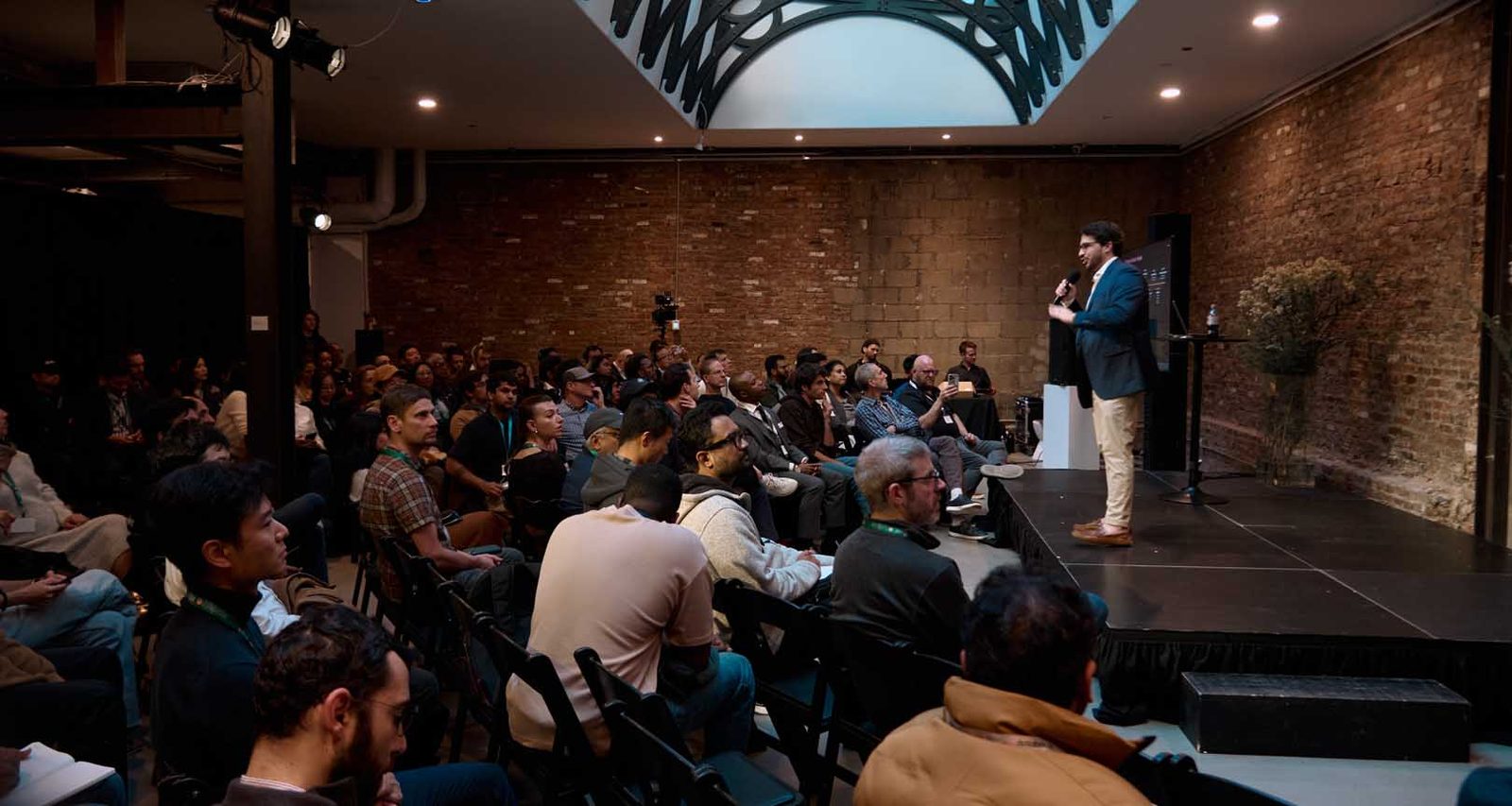 A speaker presenting to a seated audience in a brick-walled Manhattan event space, lit by stage light against a dim room